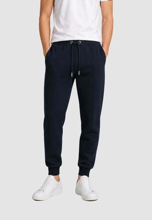 Uomo che indossa pantaloni da jogging neri con coulisse in vita e sneakers bianche, in piedi con le mani nelle tasche davanti a uno sfondo semplice.