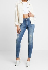 Jeans de mezclilla azul de tiro alto con acentos deslavados, combinados con una blusa blanca corta y una chaqueta de cordón color crema, llevados con mulas blancas.