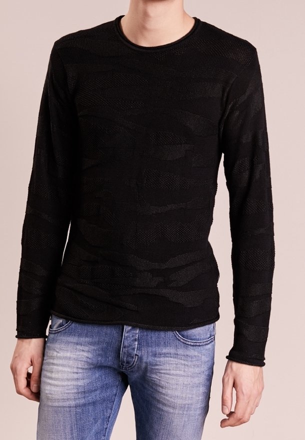 Homme portant une chemise noire texturée ajustée à manches longues associée à un jean bleu clair délavé, debout devant un fond neutre.