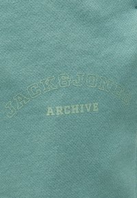 Υφασμα σε ανοιχτό πράσινο-μπλε χρώμα με αχνή κεντημένη επιγραφή "JACK & JONES" κυρτή πάνω από μεγάλα γράμματα "ARCHIVE" στο κέντρο.