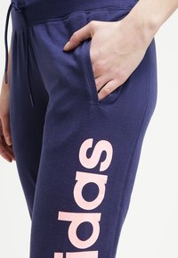 Tummansinisiä collegehousuja, joissa on teksturoitu pinta. Housuissa on sivutasku ja vasemmassa lahkeessa näkyvä pinkki "adidas" -logo.