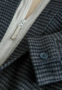 Wollmischung-Outerwear mit einem grauen Hahnentrittmuster, ausgestattet mit einem hellgrauen Reißverschluss und Knopfverschluss. Weiche Innenstruktur sichtbar.