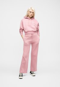 Νεαρή γυναίκα με μακριά ξανθά μαλλιά, φοράει ροζ φούτερ με κουκούλα και ταιριαστό φαρδύ παντελόνι φόρμας, μαύρα αθλητικά παπούτσια, στέκεται με τα χέρια στις τσέπες.