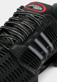 Chaussure de sport noire avec une tige en maille respirante et des accents brillants, lacets gris et une étiquette logo rouge près du col.
