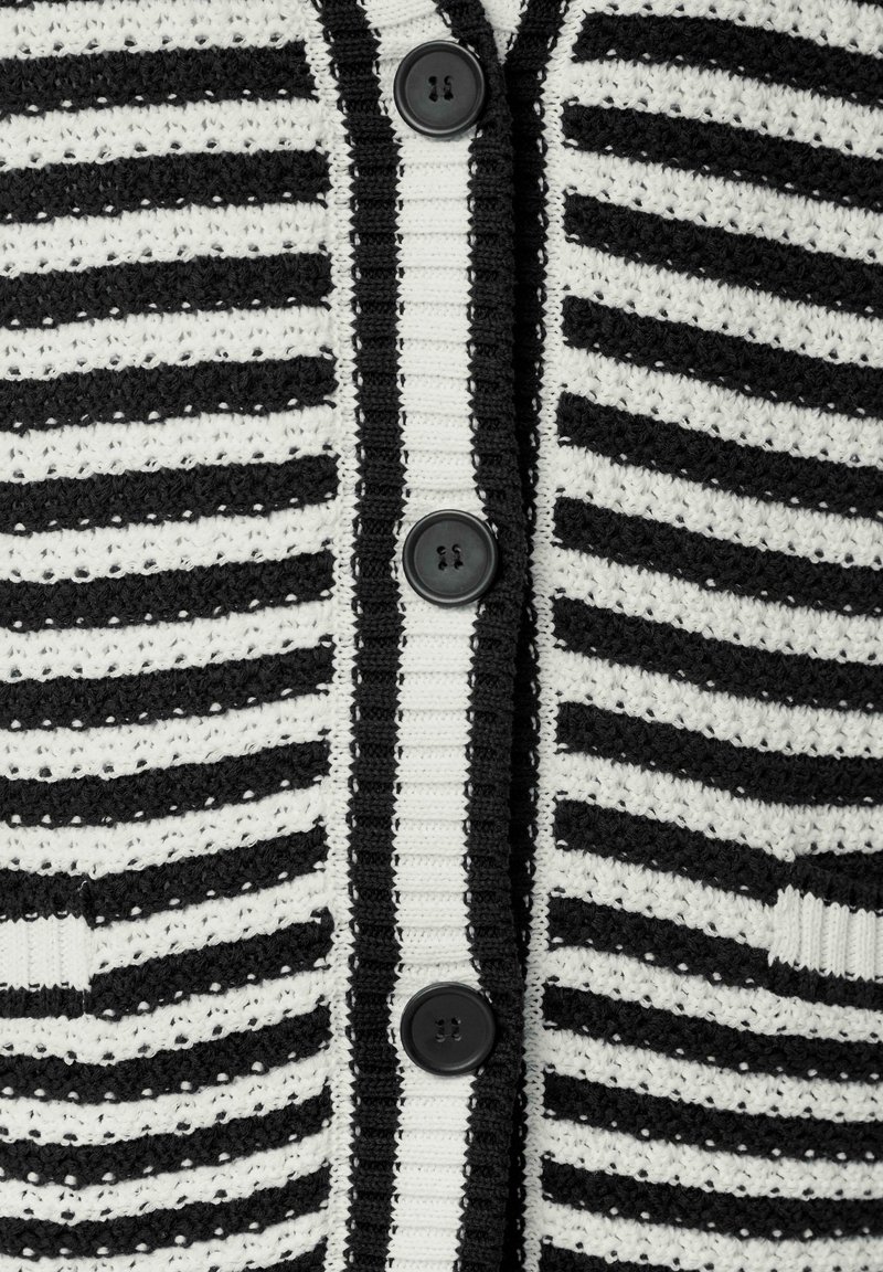 Strickjacke mit abwechselnd schwarzen und weißen horizontalen Streifen. Verfügt über fünf schwarze Knöpfe und zwei Fronttaschen.