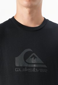 Nærbilde av en persons nakke og øvre bryst iført en svart T-skjorte med en subtil Quiksilver-logo i grått.