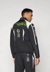 Black jacket with green and white accents, featuring logos, a high collar, and a sporty design, shown from the back.