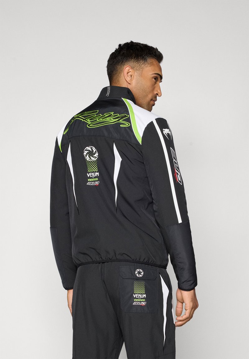 Black jacket with green and white accents, featuring logos, a high collar, and a sporty design, shown from the back.