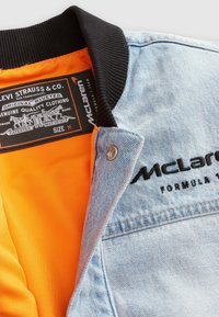 Kurtka dżinsowa z czarnym ściągaczem przy kołnierzu, pomarańczową podszewką i haftowanym logo „McLaren Formula 1”; posiada srebrny zatrzask.