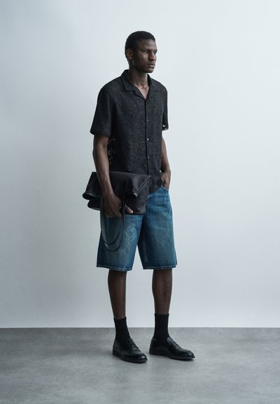 Άνδρας στέκεται φορώντας μαύρο δαντελένιο κοντομάνικο πουκάμισο, μπλε τζιν σορτς, μαύρες κάλτσες και παπούτσια, κρατώντας μια μεγάλη μαύρη δερμάτινη τσάντα.