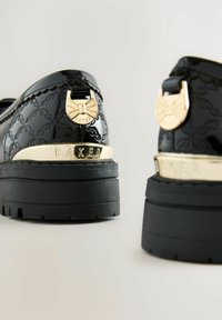 Schwarze Patelleder-Schuhe mit einer dicken schwarzen Gummisohle. Verfügt über goldene Beschläge an der Ferse, die mit "BAKER" geprägt sind, sowie ein gemustertes Design.
