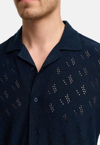 Koszula z krótkim rękawem w granatowym kolorze, wykonana z lekkiego, teksturowanego materiału z geometrycznymi wycięciami. Posiada kołnierzyk i dwa guziki przy zapięciu.