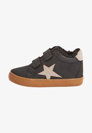 Zapato para niño pequeño negro con suela de goma marrón claro, dos tiras de velcro, parche de estrella beige y forro interior peludo.