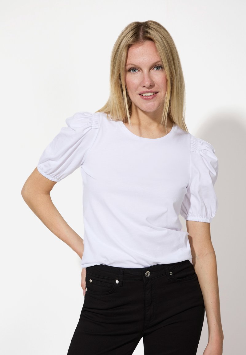 Weißes kurzärmeliges Oberteil mit puffärmel, rundem Halsausschnitt und weichem Stoff, kombiniert mit schwarzen Jeans mit Fronttaschen und Metallbeschlägen.