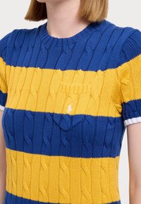 Knit short-sleeve top featuring blue and yellow horizontal stripes, cable pattern, and a small chest pocket. Trimmed with white cuffs.