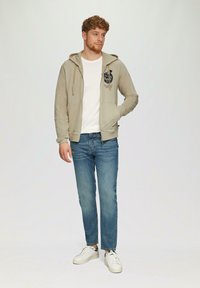 Sweat à zip beige avec poches avant, imprimé graphique, associé à un jean bleu et des baskets blanches. Tissu doux avec une coupe décontractée.