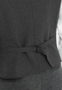 Donkergrijze op maat gemaakte vest van een soepele, gestructureerde stof met een kleine gesp en geplooide details aan de achterkant, gecombineerd met een wit shirt eronder.