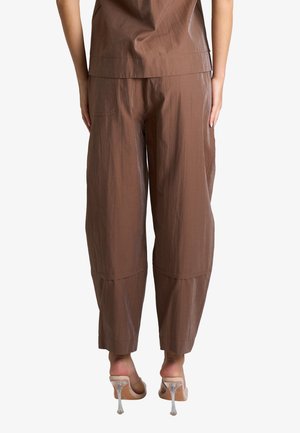 Vista posteriore di una persona che indossa pantaloni marroni larghi e un top abbinato, abbinati a sandali trasparenti con tacco su sfondo bianco.