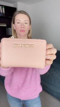 Rozā ādas maciņš ar teksturētu virsmu. Ar zelta "VALENTINO" logo un rāvējslēdzēju. Turēts rokā pret izplūdušu fonu.