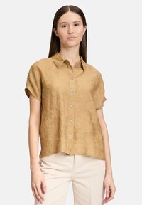 Camicia a maniche corte realizzata in tessuto beige testurizzato con un motivo sottile, dotata di colletto e apertura con bottoni, abbinata a pantaloni di colore chiaro.