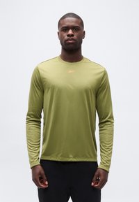 Άνδρας φοράει μακρυμάνικο αθλητικό πουκάμισο ελιάς και μαύρο παντελόνι, στέκεται μπροστά σε απλό ανοιχτό γκρι φόντο, κοιτάζοντας προς τα εμπρός.