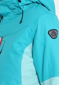 Veste à capuche turquoise et bleu clair avec des cordons noirs et un patch logo circulaire noir et blanc sur la partie supérieure de la manche.