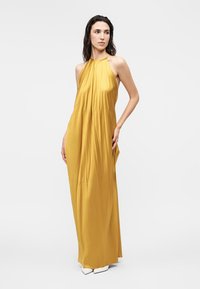 Γυναίκα που φοράει χωρίς μανίκια μακριό φόρεμα χρώματος μουστάρδας με halter neck και λευκά μυτερά παπούτσια, στέκεται μπροστά σε απλό φωτεινό φόντο.