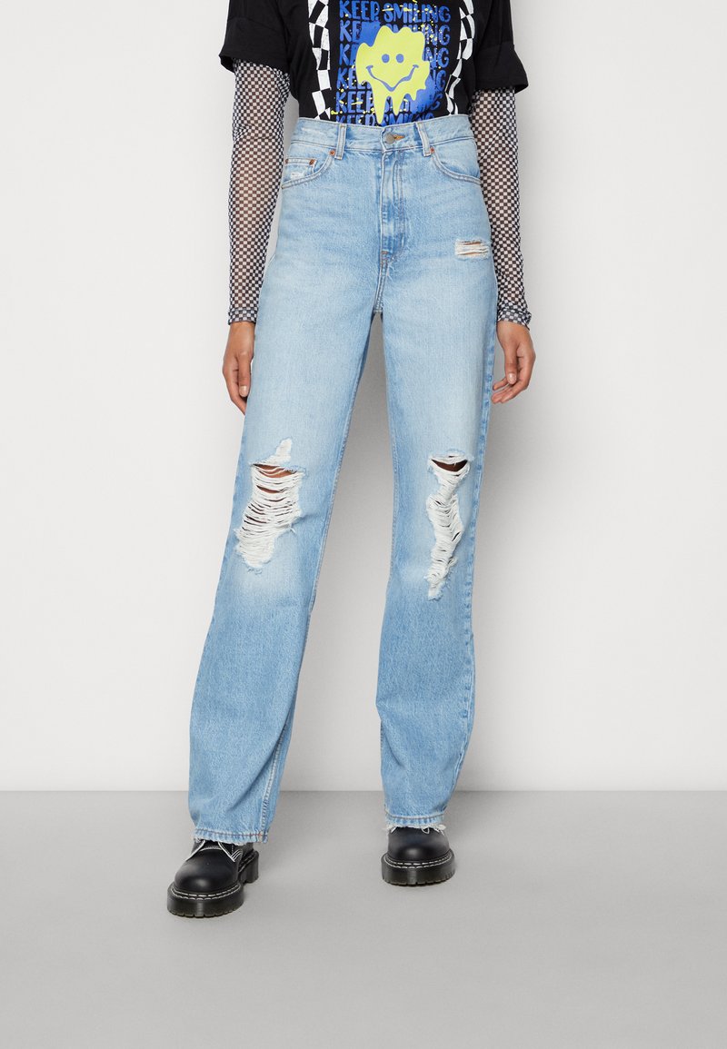 Jeans de cintura alta azul claro, com um corte solto, apresentando rasgos proeminentes nos joelhos e uma bainha desfiada. Combinados com uma t-shirt gráfica preta.