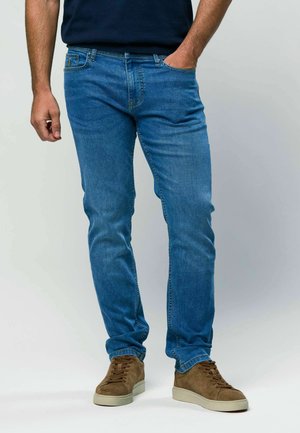 Mężczyzna w niebieskich dżinsach i brązowych zamszowych sneakersach stoi z jedną ręką w kieszeni na jednolitym tle.