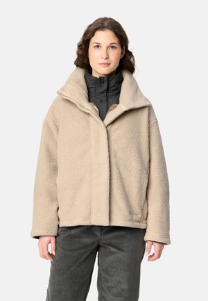 Vrouw draagt een beige teddyjas over een zwarte jas, gecombineerd met donkergrijze broek, staand tegen een effen witte achtergrond.