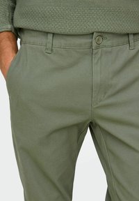 Chino en coton vert olive avec une texture lisse, comportant des poches latérales et une fermeture à bouton à la taille. Design classique à jambe droite.