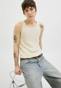 Canotta bianca a costine con scollatura rotonda, indossata con jeans in denim larghi di colore blu chiaro e una cintura marrone, con tatuaggi sulle braccia.