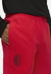 Røde sweatpants med elastisk talje, sidelommer og et broderet sort logo med teksten "ACM 1899" på venstre lår. Blød stoftekstur.