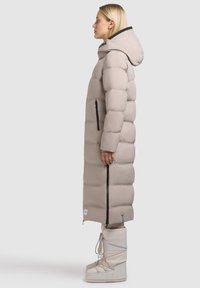 Vrouw in een lange beige donzen jas met capuchon en beige winterlaarzen, staande in profiel tegen een effen achtergrond.