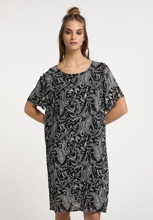 Nuori nainen vaaleanruskeilla hiuksilla puolikampauksessa, musta mekko jossa valkoiset monimutkaiset paisley- ja kukkakuviot, seisoo kädet selän takana.