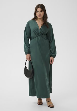 Femme debout portant une longue robe en satin vert foncé à manches longues, tenant un sac à main noir, et chaussée de talons noirs à brides.