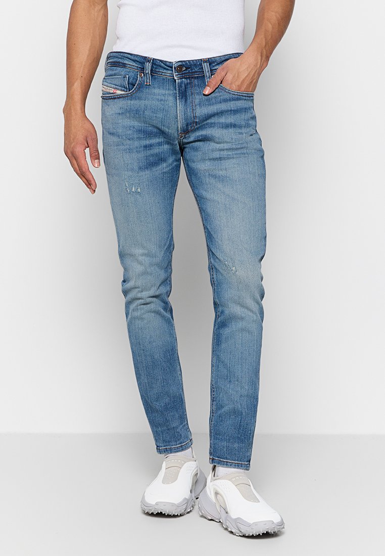 Homme portant un jean slim-fit bleu clair avec une légère déchirure, des baskets blanches et un t-shirt sans manches blanc, se tenant devant un fond uni.