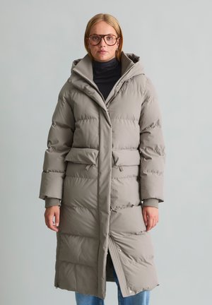 Woman wearing long beige puffer coat with hood, black turtleneck, blue jeans, and large brown glasses standing against a plain light background.