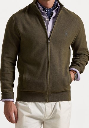 Homme portant un pull zippé vert olive sur une chemise violet clair et un foulard bleu marine à pois, avec un pantalon blanc et une montre-bracelet.