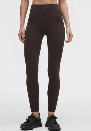 Vrouw draagt donkerbruine high-waisted legging en zwarte sportschoenen, staand tegen een effen lichte achtergrond.