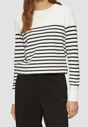 Kvinde iført en hvid sweater med sorte vandrette striber og sorte bukser, vist fra skuldre til hofter mod en grå baggrund.