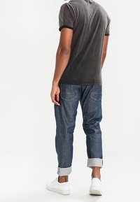 Grå t-shirt, korta ärmar, vy bakifrån. Blå denimjeans, uppvikta fållar, två bakfickor. Vita sneakers med texturerade sulor.