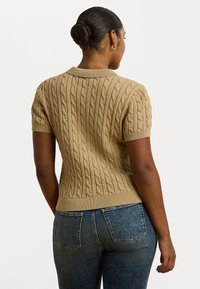 Pull en tricot beige à manches courtes avec motif en cable et ourlet côtelé. Porté avec un jean bleu, montrant la vue arrière et le design du col rond.