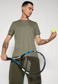 Oliven grønn idretts t-skjorte med ribbet tekstur og logoakcent, kombinert med matchende shorts og holder en tennisracket og en neongul tennisball.
