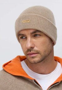 Beige knitted beanie with ribbed texture and a leather brand label in the front. Worn with a beige hoodie and orange accents at the collar.