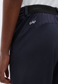 Κοντινό πλάνο ενός ατόμου που φοράει ναυτικό μπλε σορτς με το κείμενο "Daily" κεντημένο κοντά στη μέση και μια μαύρη ελαστική ζώνη.