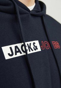 Sötétkék kapucnis pulóver, fehér téglalap alakú logóval, amelyen a "JACK&" feketével és a "JONES" vörössel szerepel. Vastag húzózsinórok és puha textúra.