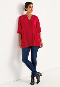 Rode tuniek met een V-hals en knoopdetails, driekwartmouwen, textuurstof, gecombineerd met blauwe skinny jeans en zwarte enkelboots.