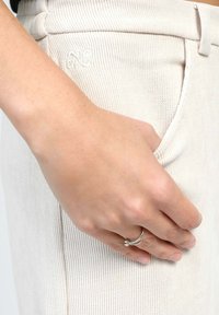 Pantalons beige à côtes avec une coupe ajustée. Une main repose sur la poche, mettant en valeur une bague en argent aux doigts. Broderie discrète sur la ceinture.