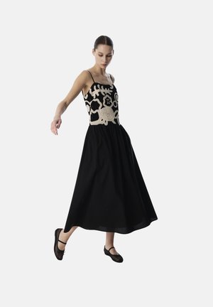 Abito lungo nero con design senza maniche, caratterizzato da un intricato ricamo beige e una gonna svasata. Modella che indossa ballerine nere.